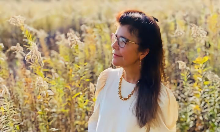 woman with long dark hair and glasses in a white dress and thick gold necklace in a field of goldenrod