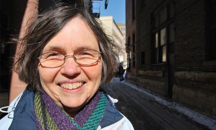 Margaret Rozga wearing a scarf and glasses smiling in front of an alley lined by brick buildings and a dusting of snow