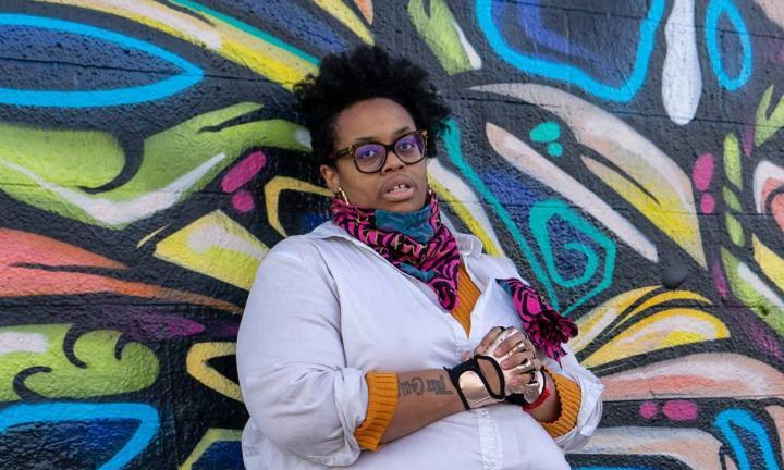 author Naima Lowe standing with clasped hands in front of colorful graffiti