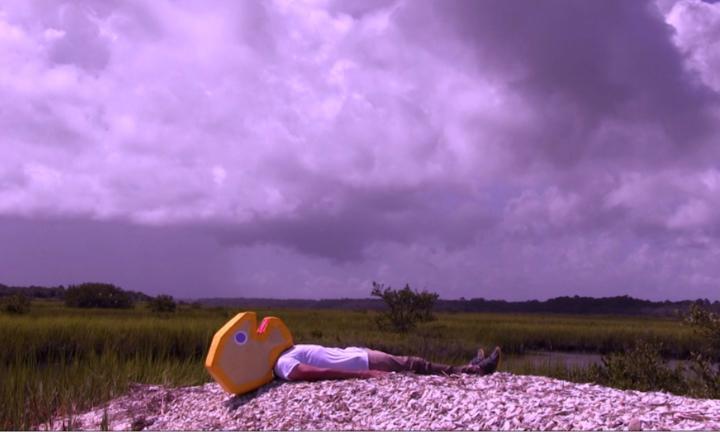 Man on a pile of mulch wearing a large round cardboard head with a yellow outline under a gray cloudy sky and with a green far horizon