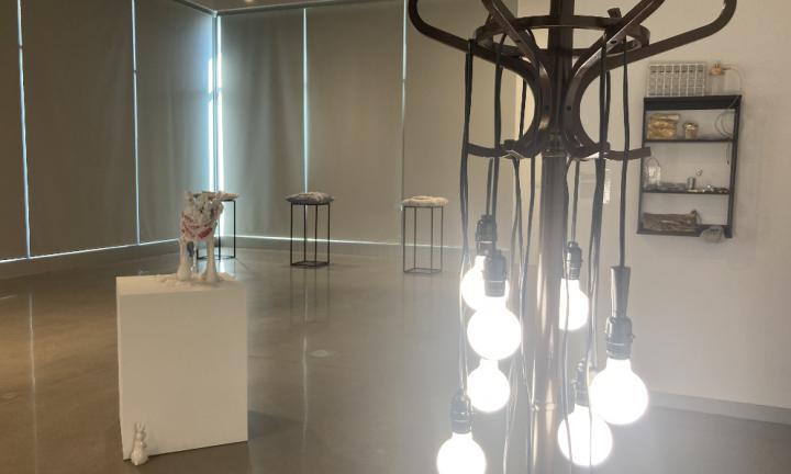 gallery space with white pedestal and ceramic figures, curved coat rack with several hanging lit Edison bulbs