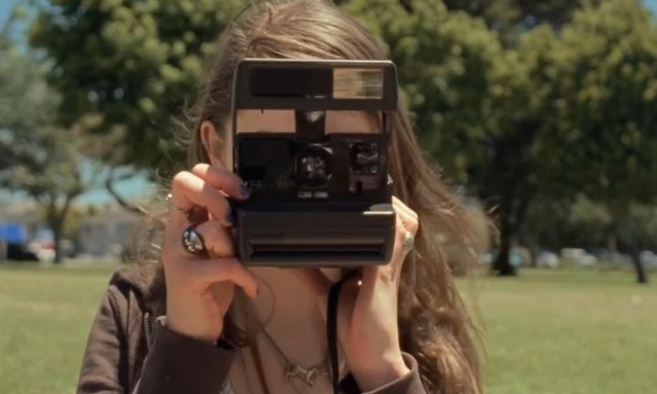 woman with long hair standing in front of trees with her face obscured by a large black Polaroid camera