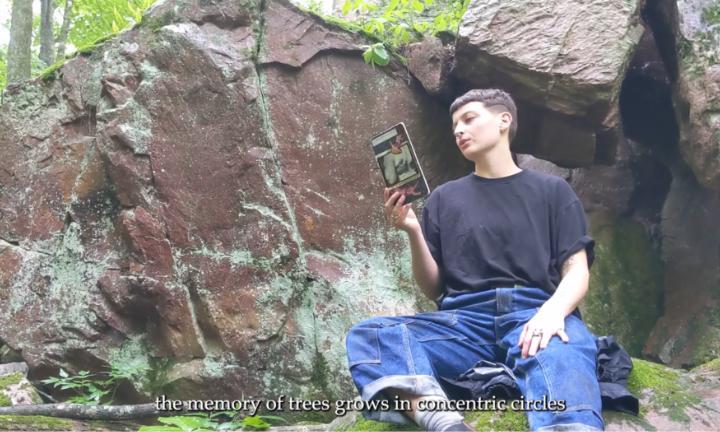 poet in black t-shirt and blue jeans reading a journal sitting on a boulder with greenery
