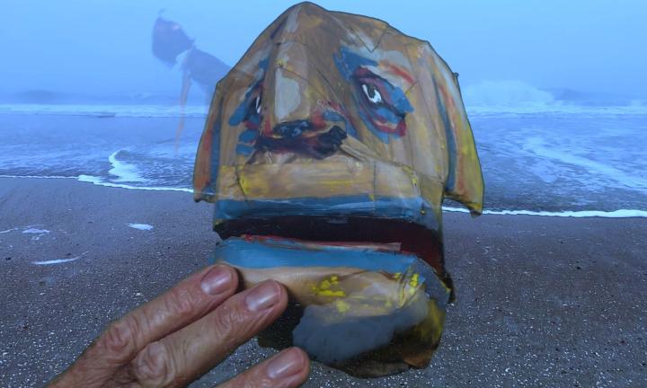 large dog puppet head with a human hand at its chin superimposed over a beach