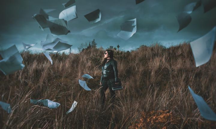 woman carrying a typewriter through a field with papers flying all around under a cloudy sky