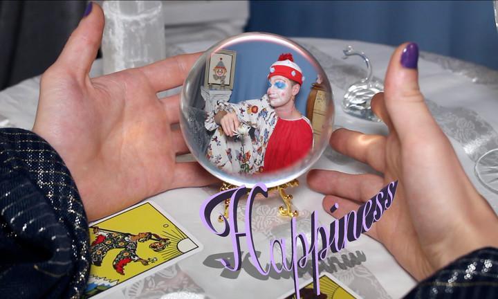 hands caressing a crystal ball with a clown in face paint and a dotted red and white hat depicted within it. The word "happiness" connects the crystal ball to the palm of the right hand