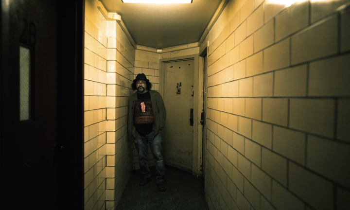 Photo of man at end of shadowy building hallway. Door behind him reads 2A. 