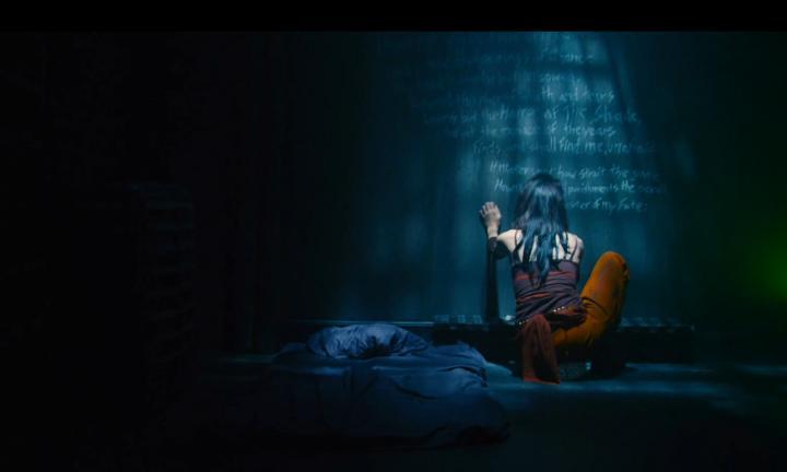 person with long dark hair squatting on a floor writing on the black wall in chalk
