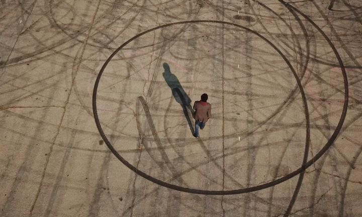 Aerial shot of a man walking across pavement marked with large arcs and circles. His shadow is to the top left of him