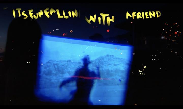 yellow words "its fun fallin with a friend" handwritten about a blue tiled screen of a shadowy figure wearing a hat in front of mountains