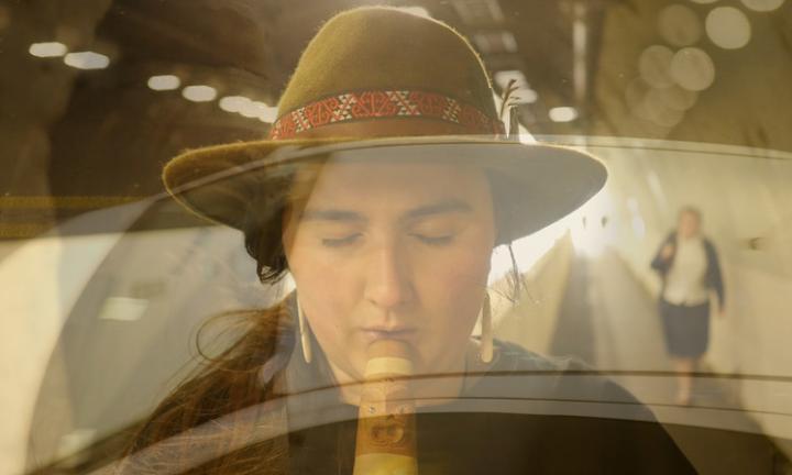 woman in felt hat with beaded band playing a wind instrument with a lighted tunnel and a pedestrian walking toward camera