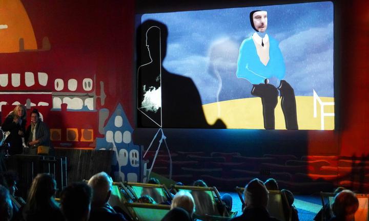 audience looking at a large screen of a collage man with a real face seated talking to a shadow figure with its back to the camera