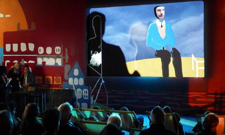 collage image of a seated man talking to a shadowed figure with their back to the viewer projected on a screen with audience looking up at it