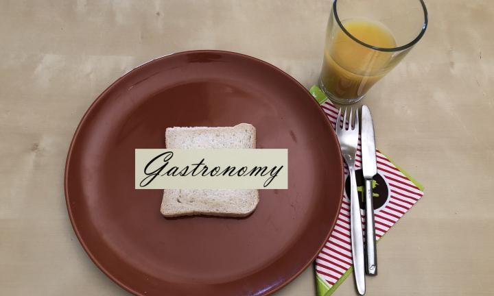 maroon plate with slice of bread and the word "gastronomy" laid across it. with a glass of orange juice and a fork and a knife resting on a striped napkin beside the plate