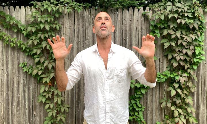 man with his two hands raised in front of a fence with vines