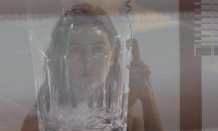 young woman with long hair and bare shoulders holding a corded phone upside down away from her face behind a beveled drinking glass that obscures her face