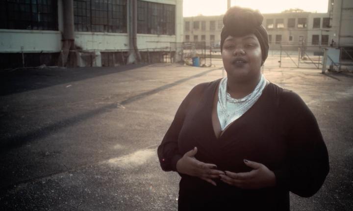 Black woman with a dark head wrap on and black dress with silver necklaces hands cupped in front of her standing in a parking lot with industrial buildings in the background