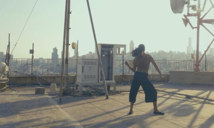 Figure dancing on a roof crowded with towers and equipment against a city skyline