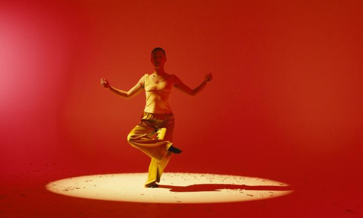 young woman with hands raised standing on one leg in front of a reddish orange background
