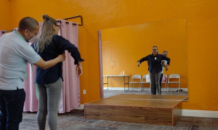 a woman holding our her arms in front of a mirror in an orange room with a man measuring her