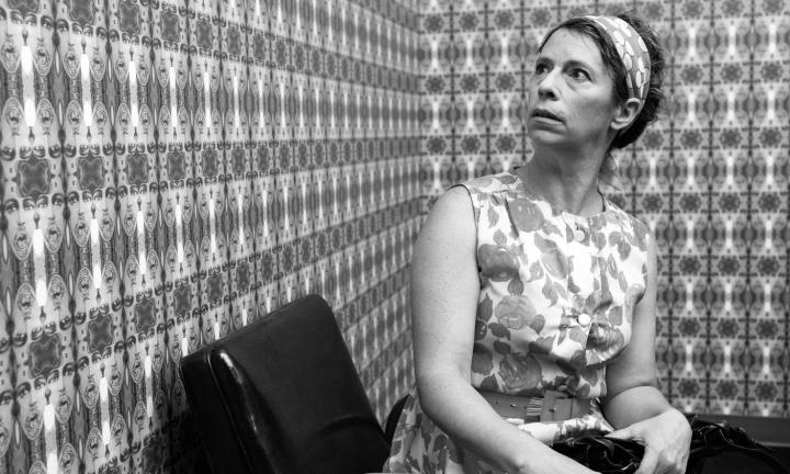 black and white photo of woman in floral dress, seated in chair, and looking up at patterned wallpaper 