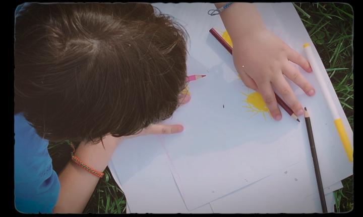 picture of the back of a child resting on the grass drawing a sun with colored pencils