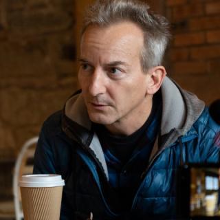 seated man with gray hair wearing a blue puff jacket looking to the side with a disposable coffee cup in front of him
