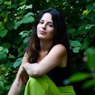 Photo of author wearing black and green, crouching in front of a green backdrop of foliage 