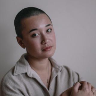 Poet Megan Kim with shaved head and tan button-up shirt against a grey background