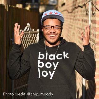 smiling young Black man wearing glasses, blue cap with stars, and a black hoodie with words "black boy joy" in white 