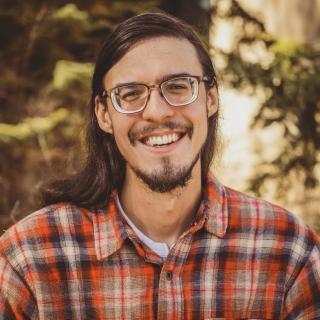 smiling man with long hair and glasses wearing a grey and orange plaid shirt
