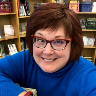 Author posing in front of bookshelves, smiling and wearing red glasses.