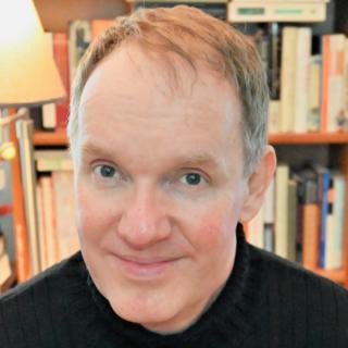 man with dark eyes wearing a black turtleneck smiling in front of bookshelves